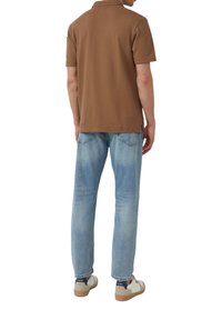 Braunes Poloshirt mit kurzen Ärmeln, kombiniert mit hellblauen Denim-Jeans und weißen Sneakers mit navyfarbenen Akzenten. Einfaches Design, lockere Passform.