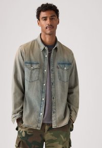 Άνδρας με κοντά σγουρά μαλλιά και μουστάκι, φορεί ξεθωριασμένο τζιν πουκάμισο πάνω από γκρι μπλουζάκι και παραλλαγές παντελόνια cargo, με τα χέρια στις τσέπες.