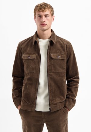 Bruine corduroy jas met een kraag, twee borstzakken en een ritsluiting. Draag deze over een lichtgekleurde geribde trui.