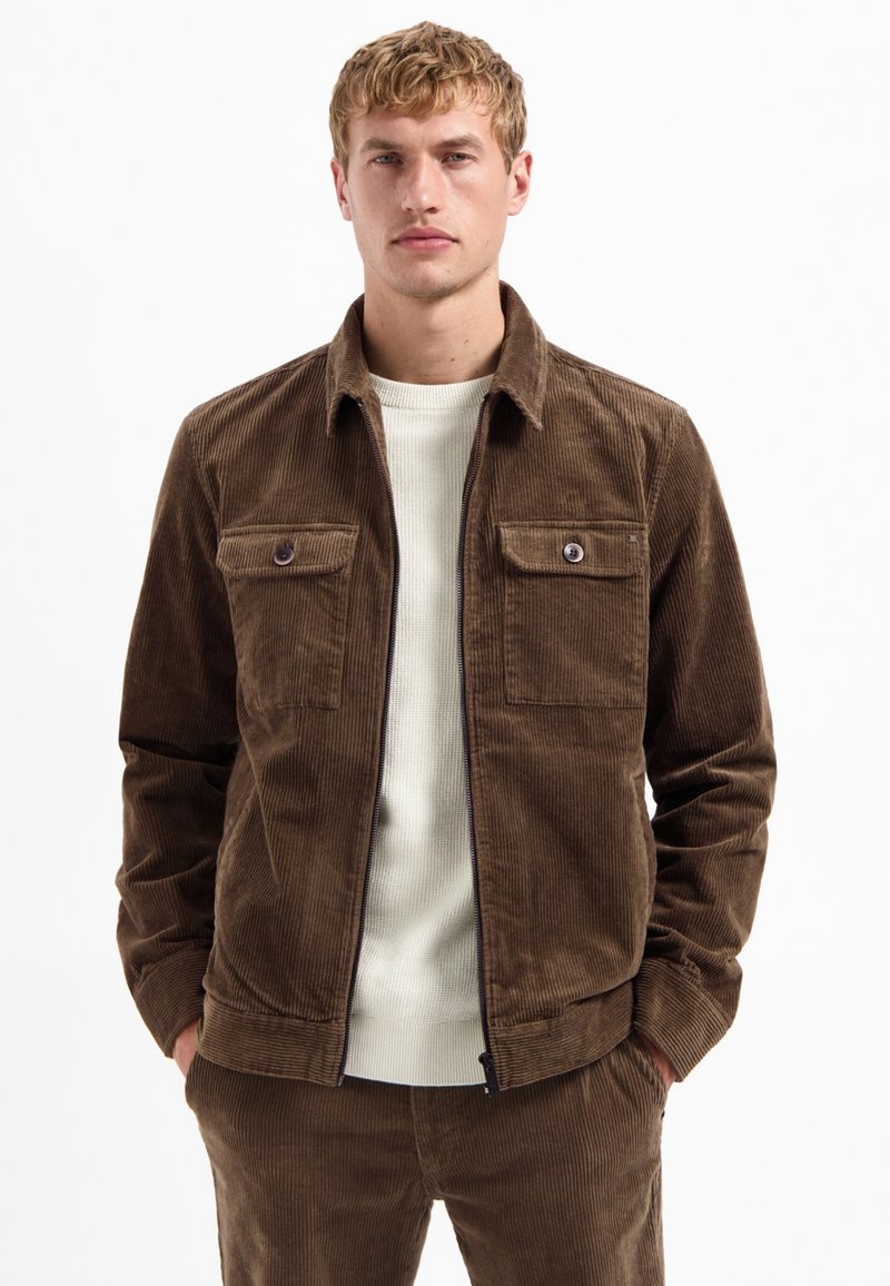 Bruine corduroy jas met een kraag, twee borstzakken en een ritsluiting. Draag deze over een lichtgekleurde geribde trui.