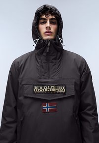 Jachetă pulover într-o nuanță închisă de gri, cu guler înalt, buzunar frontal, logo "Napapijri" și o aplică cu steagul Norvegiei. Dispune de glugă cu șnur și fermoare.