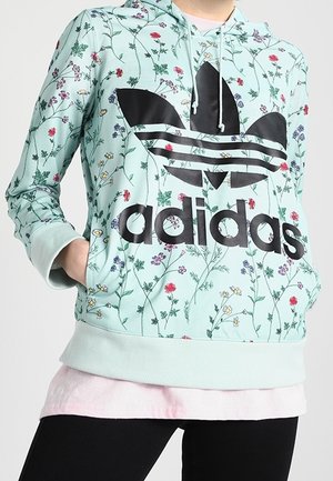 Osoba nosząca jasnoniebieską, kwiatową bluzę z kapturem Adidas z czarnym logo i kieszenią kangura, w połączeniu z czarnymi spodniami i jasnoróżową podkoszulką.