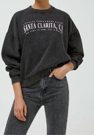 Osoba nosí vypranou černou mikinu s nápisem "Santa Clarita, CA" ve spojení s vysokým pasem tmavě šedých džín, jedna ruka v kapse.