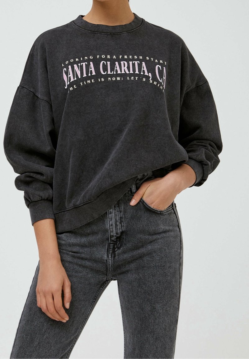 Personne portant un sweat-shirt noir délavé avec le texte « Santa Clarita, CA » associé à un jean gris foncé taille haute, une main dans la poche.