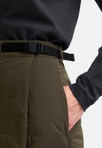 Olijfgroene gewatteerde broek met een gestructureerde afwerking, voorzien van een zwarte verstelbare riem en zijzakken. Zwart langslievend shirt eronder.