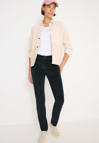 Světle béžová kordurová košile-jacket, bílý tričko, tmavě zelené kordurové kalhoty a krémové sneakers. Růžová čepice na hlavě, postoj stojící.