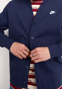 En person knäpper en marinblå Nike cardigan över en röd- och vitrandig skjorta, med silverringar på båda händernas.