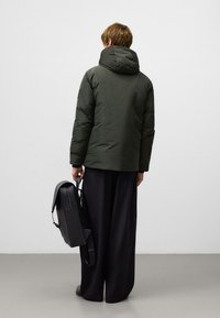 Oliwkowa kurtka puchowa z wysokim kołnierzem, noszona na luźnych czarnych spodniach. Osoba trzyma czarny skórzany plecak, łączący matowe i gładkie tekstury.