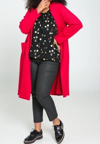 Cardigan long rouge porté sur un chemisier noir à imprimé floral avec des accents dorés, associé à un pantalon noir ajusté et des chaussures noires brillantes.