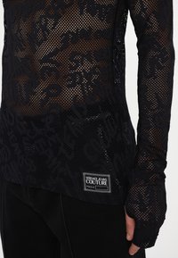 Čierny sieťovaný top s dlhými rukávmi, ktorý obsahuje textúrované písmená, okrúhly výstrih a výraznú nášivku loga Versace Jeans Couture na leme.