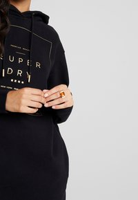 Kvinna som bär en svart hoodie med guldtexten "Superdry" och håller samman sina händer, bär en stor orange ädelstenring på sitt finger.
