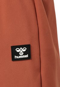 Oranje stof met een gladde textuur, voorzien van een zwart label met een bij-logo en de tekst "hummel" prominent weergegeven.