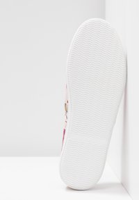 Semelle en caoutchouc blanche d'une chaussure de sport avec un motif de bande de roulement ondulé, positionnée à la verticale contre un mur d'angle blanc sur une surface blanche.