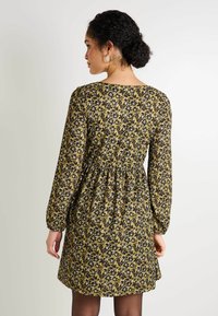 Vestido floral en amarillo y púrpura oscuro, con mangas largas abullonadas y una cintura fruncida, con un dobladillo a la altura de la rodilla y tela texturizada.