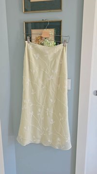 Lysegul nederdel lavet af gennemsigtigt stof, med hvid blomsterbroderi og en flydende design. Hængende på en metalclip med en træstang.