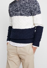 Marinblå och vit stickad tröja med ett strukturerat kabelmönster, ribbade ärmar och nederkant. Bärs över en vit skjorta med ett svart mönster.