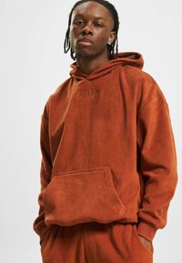 Fleece hoodie v barvni rust z sprednjim žepom, kapuco s vezalko in subtilnim embosiranim logotipom na prsih. Mehka tekstura, sproščen kroj.