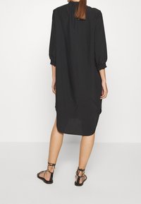 Femme aux longs cheveux portant une robe noire ample jusqu'aux genoux avec des manches 3/4 et des sandales plates noires à lacets, se tenant devant un fond blanc.