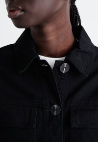 Musta farkkutakki, jossa on kaulus, kaksi etutaskua ja yksinkertainen nappisuljenta, tekstuuripintaisena.