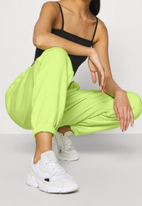 Person som sitter på huk och bär neongröna joggingbyxor, vita Adidas-sneakers och en svart ärmlös topp mot en enkel ljus bakgrund.