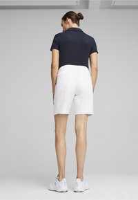 Pantalones cortos blancos con una textura suave y una cinturilla elástica, combinados con una polo azul marino. Usados con zapatillas deportivas blancas. Vista trasera.