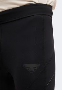 Gros plan sur un legging de sport noir avec un logo triangulaire discret sur la partie supérieure de la cuisse et des coutures visibles.
