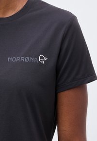 Κοντινό πλάνο σκουρόχρωμης γκρις μπλούζας με το κείμενο "NORRØNA" και λογότυπο κράνους Βίκινγκ στο στήθος, που φοράει άτομο με μεσαίο καφέ δέρμα.