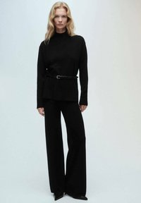 Crna pletena bluza s visokim izrezom, uparena s crnim hlačama širokih nogavica. Crni remen naglašava struk. Cijela kombinacija ima glatku teksturu.