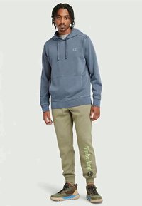 Zils kokvilnas kapučinsponsors ar priekšējo kabatu, saskaņots ar olīvu zaļām sporta biksēm, uz kurām ir uzraksts "Timberland" zaļā krāsā. Kārtīgi brūni un pelēki sporta apavi noslēdz šo tērpu.