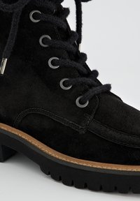 Bottine en daim noir avec semelle en caoutchouc, œillets en métal et lacets épais, dotée d'une finition texturée et d'un détail de semelle marron en contraste.