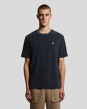 Ung mann iført teksturert mørk marineblå t-skjorte med liten gul fuglelogo på brystet og beige cargobukser, stående mot ensfarget lys grå bakgrunn.