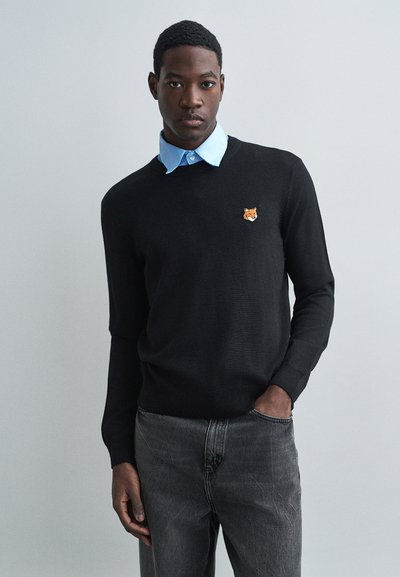 Giovane uomo che indossa un maglione nero con logo di volpe sopra una camicia color azzurro chiaro, jeans grigio scuro, in piedi contro uno sfondo grigio chiaro semplice.