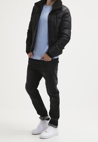 Uomo che indossa un giubbotto imbottito nero con colletto in pelliccia, un maglione azzurro chiaro, jeans neri arrotolati alle caviglie e sneakers bianche, in piedi con le mani nelle tasche.