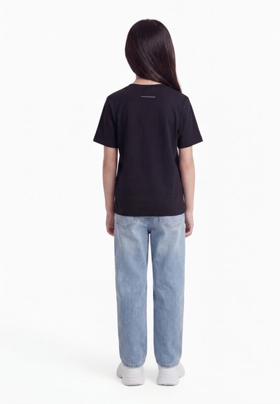 Kind met lang donker haar staat met de rug naar voren, draagt een zwart T-shirt, lichtblauwe spijkerbroeken en witte sneakers, tegen een witte achtergrond.