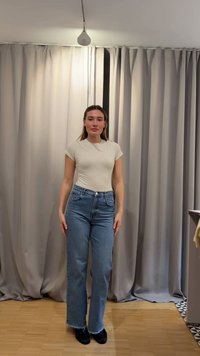 Mujer de pie en el interior, con una camiseta beige y jeans azul de talle alto, frente a unas cortinas beige cerradas sobre un suelo de madera.