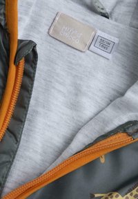 Grå huvjacka med orange dragkedja, med en mjuk textur och ljusgrått tyg, märkt med "My First Chicco"-etikett.