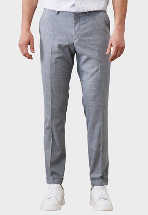 Mand iført lysegrå, slim-fit bukser med diskret ternet mønster og hvide sneakers, stående mod en ensfarvet baggrund.