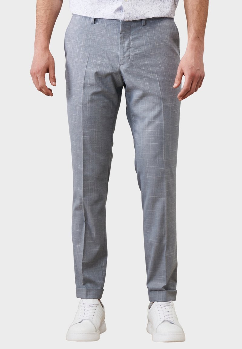 Mężczyzna w jasnoszarych dopasowanych spodniach z subtelnym wzorem w kratę i białych trampkach, stojący na jednolitym tle.