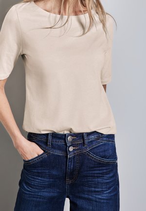 Mujer vistiendo una camisa de manga corta beige metida dentro de unos jeans de doble botón azul oscuro, con una mano en el bolsillo, contra un fondo neutro.