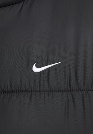 Melnā pūkainā jaka ar izšūtu dizainu un baltu izšūtu Nike logotipu kreisajā pusē. Izgatavota no gludas sintētiskas materiāla.