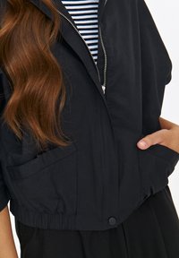 Czarna, krótka kurtka z zamkiem z przodu, elastycznym dołem i dwiema kieszeniami. Posiada krótkie rękawy i gładką fakturę. Pod spódkiem znajduje się koszula w paski.