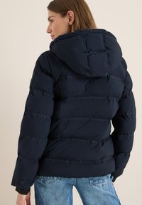 Marinblå pufferjacka med huva, som har en quiltad design och synliga sömmar. Utformad med långa ärmar och en avslappnad passform.