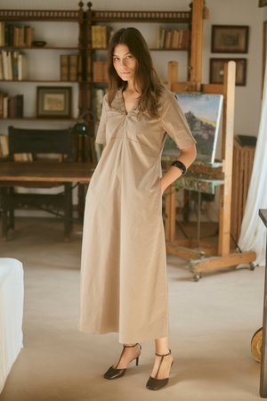 Femme en robe beige longue jusqu'aux chevilles et talons noirs, debout dans une pièce confortable avec un chevalet en bois et des étagères remplies de livres.