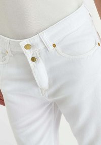 Witte spijkerbroek van denim met gouden knopen en klinknagels, gedragen met een wit geribd topje, waarbij details van het voorvak en de tailleband zichtbaar zijn.