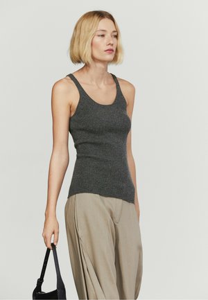 Donna con capelli biondi a caschetto che indossa un top senza maniche in maglia grigio scuro e pantaloni beige larghi, tiene una piccola borsa nera nella mano destra.