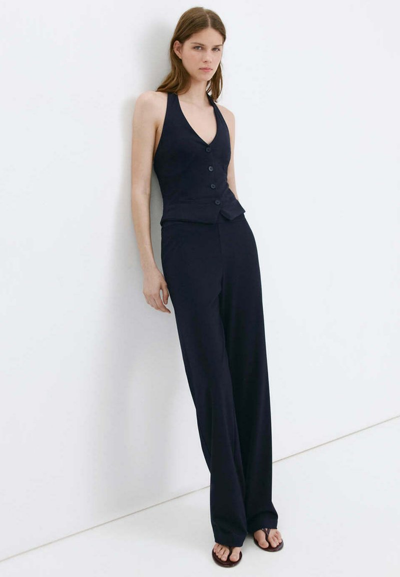 Jeune femme appuyée contre un mur blanc, portant un gilet noir sans manches boutonné, un pantalon large et des tongs noires.