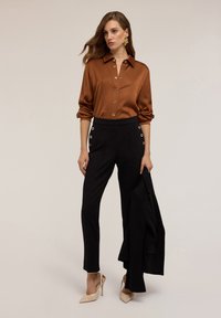 Braune Satin-Hemd mit Knöpfen und hochgeschlagenen Ärmeln, kombiniert mit schwarzen Hosen mit seitlichen Ösen und nackten High Heels.