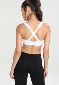 Top deportivo blanco con tirantes cruzados, que presenta acentos de herrajes circulares y una textura suave, combinado con legging ajustados negros.