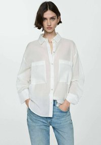 Camicia bianca semi-trasparente con due tasche sul petto, vestibilità rilassata e maniche arrotolate, abbinata a jeans azzurri chiari.
