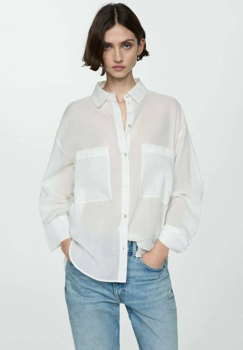 Camicia bianca semi-trasparente con due tasche sul petto, vestibilità rilassata e maniche arrotolate, abbinata a jeans azzurri chiari.